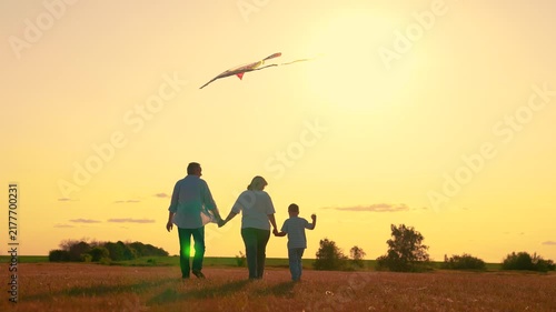 Family plays with kite in sky, picnic in field. Happy family with child plays with kite in park. Family walks on lawn, child dreams flying. People travel together, nature, silhouette. Child wants fly