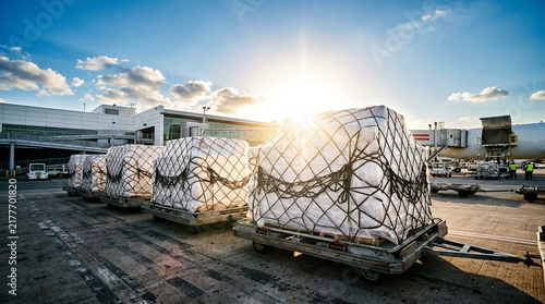 Air cargo pallets wrapped in protective plastic and cargo nets on transport trailers at an airport apron during sunset with a terminal building and aircraft in the background.