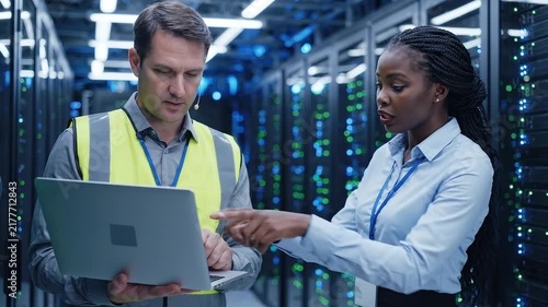 Data center technicians collaborate while reviewing server infrastructure on a laptop in a high-tech facility.