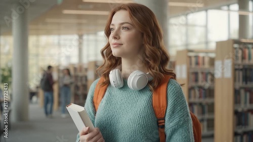 Student with wireless earbuds and backpack walks through a modern library while holding a smartphone.
