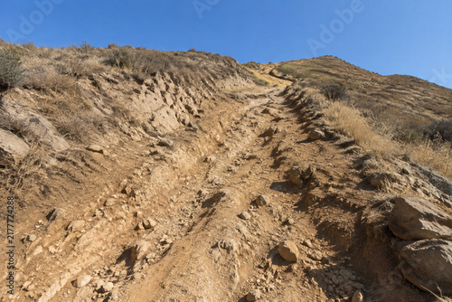 Steep unpaved trail on dry rocky hillside with ruts and exposed roots, arduous hiking path concept