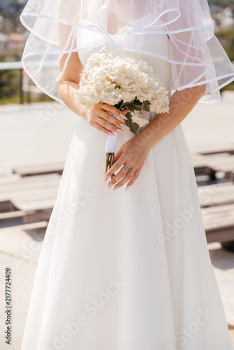 A young Caucasian woman in a white wedding dress holds a bouquet of white flowers. She wears a sheer veil and stands outdoors in a bright setting.