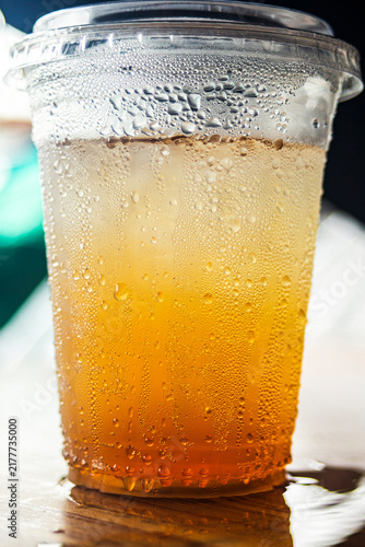 Iced amber beverage in a plastic cup with condensation on a wooden table.A refreshing eye-level shot of a cold amber-colored drink, such as iced tea, apple juice, or traditional herbal beverage