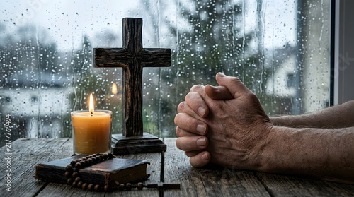 Elderly person praying with hands clasped by a wooden cross and candle on a rainy day background for spiritual reflection