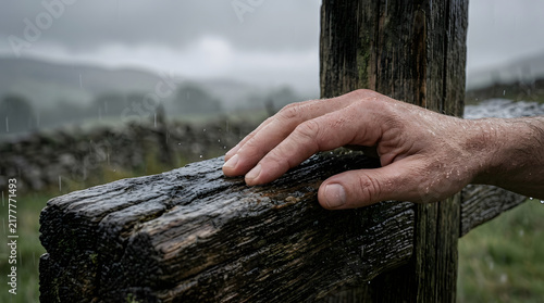 A person hand resting gently on a weathered wooden cross outdoors in the rain against a scenic misty green countryside landscape background
