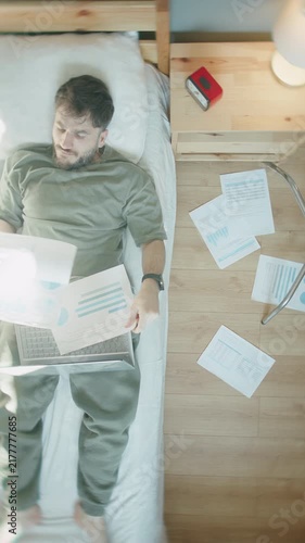 Man working in bed with laptop and scattered charts, papers falling around ,vertical video