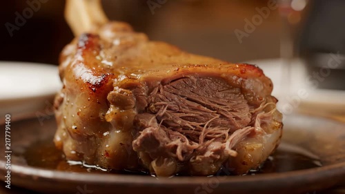 Close-up of a tender, slow-cooked meat shank, possibly lamb or beef ossobuco, with rich brown gravy served on a rustic wooden plate. High-quality food photography.