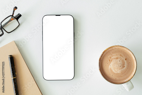 Top view flat lay of modern smartphone mockup with blank white screen on clean desk alongside coffee cup, notebook, pen, and eyeglasses. Minimalist workspace template for design.