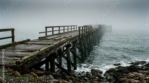 A weathered wooden pier extends into a misty ocean. The pier’s planks are gray and worn, showing signs of age and exposure to the elements.
