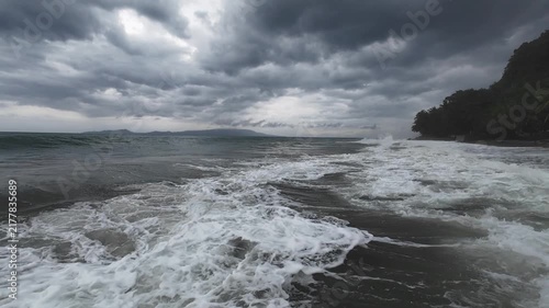Stormy ocean waves crash onto a tropical shoreline under heavy clouds. Cinematic wide footage capturing the raw power of nature and the dramatic moody atmosphere.