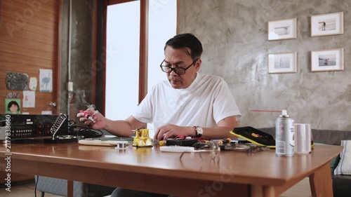 Mature Asian man concentrates on repairing home electrical appliance, disassembling and soldering circuit board, tired from precision detail focusing, taking a break, resting from his workshop hobby.
