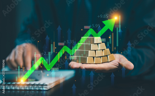 Businessman holding stack of gold bullion bars with green rising stock graph and candlestick chart overlay, gold investment and wealth concept.