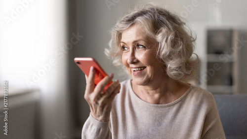 Fotografía Smiling senior woman holding smartphone, sending voice messages, speaking with virtual assistant, or talking on speakerphone at home