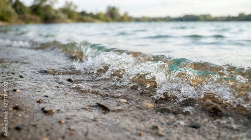 Close up of gentle waves lapping on a sandy shore with blurred background of water and trees.