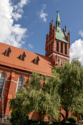  Red brick church tower with a tall green spire rising above a tiled rooftop against a dramatic blue sky with white clouds, detailed historic architecture captured from a low angle