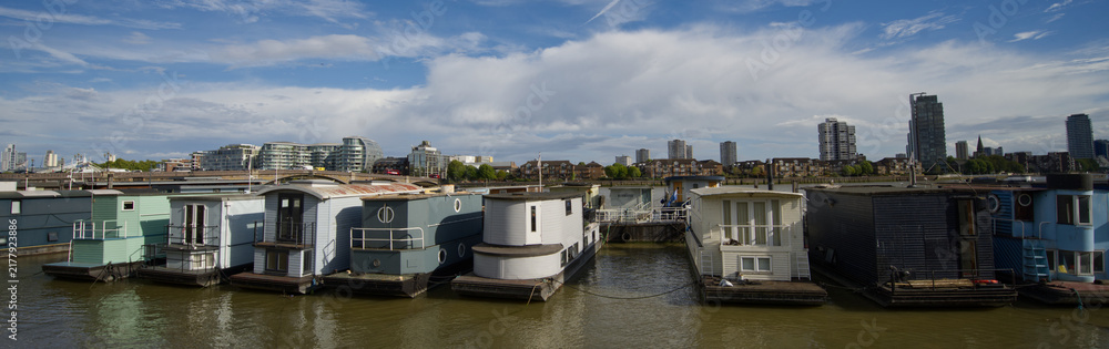 Obraz premium Colorful residential houseboats moored along the River Thames in London, England, with modern high-rise buildings and the city skyline visible in the background.