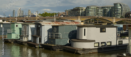 Photography Colorful residential houseboats moored along the River Thames in London, England, with modern high-rise buildings and the city skyline visible in the background