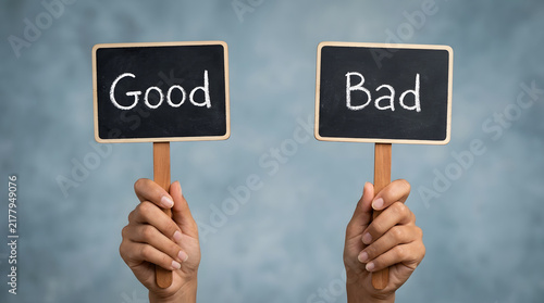Two hands holding small chalkboard signs with Good and Bad written on them against a light blue gradient background featuring comparison, choice, decision, contrast, positive, negative and evaluation