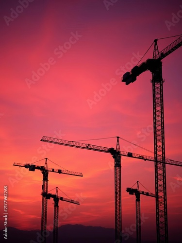 Silhouettes of construction cranes against a vibrant orange-pink evening sky, with wispy clouds drifting across the horizon, structure, || cityscape