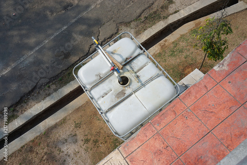 Fotografía A high-angle shot shows a large white plastic IBC water tank encased in a metal cage, placed on the ground next to a tiled patio and asphalt driveway