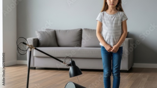 Sad girl standing near broken lamp at home