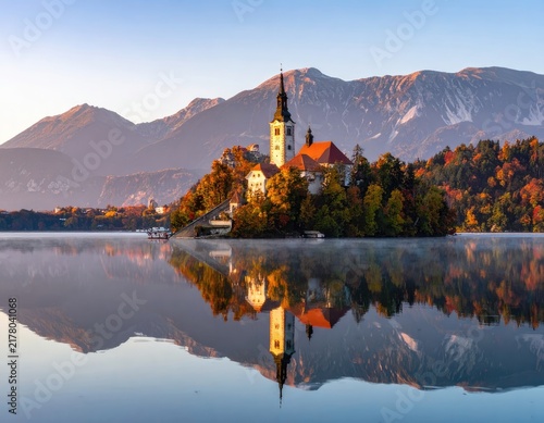 A picturesque island church with a bell tower is reflected in a tranquil lake surrounded by autumn foliage and mountains.