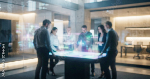 A diverse group of business professionals collaborating at a high-tech interactive table in a modern office space. Innovative technology enhances teamwork and creativity.