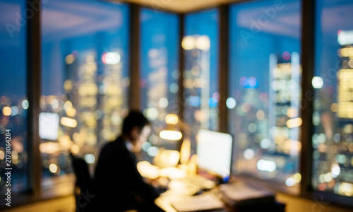 A blurred interior view of an office at night, showcasing a person working at a desk with city lights illuminating the skyline through large windows.