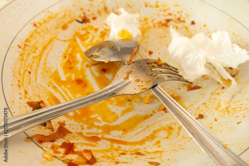 Dirty white plate smeared with orange sauce, used fork and spoon crossed over food remnants and crumpled napkin, conveying after-meal mess, leftovers and everyday dining cleanup moments.