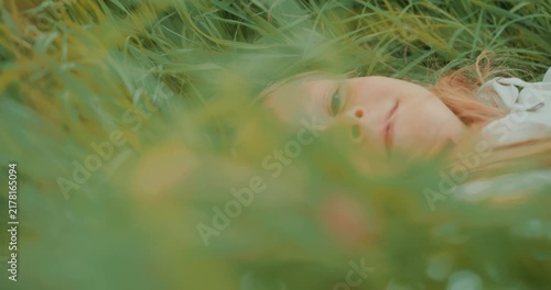 Cinematic side view of a young girl lying in tall green grass. She slowly turns her head and looks directly at the camera through the grass. Natural light, shallow depth of field. High quality 4k