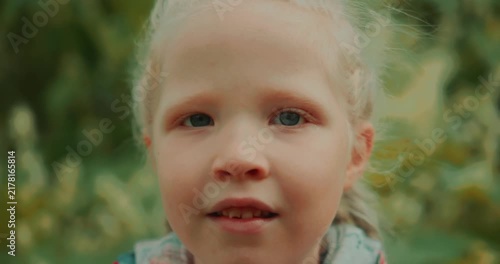 Close-up portrait of a young girl looking directly at the camera outdoors. At the end of the shot, she gives a slight smile. Natural light, green background, static camera. High quality 4k footage