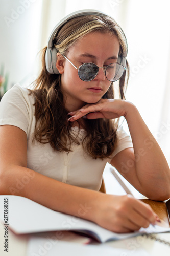 A teenage girl with strabismus wearing special corrective eyeglasses with a lens patch studies at a desk. She wears headphones while writing in a notebook during school preparation.