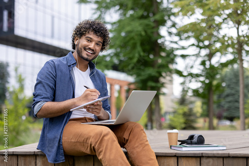 Young mixed-race student with curly hair and earphones sits on a campus bench  smiling while writing in a notebook  laptop and coffee nearby for remote study and work