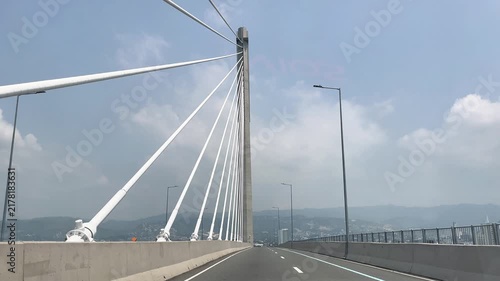 Driving across a modern cable stayed bridge along a modern highway expressway with white cables and concrete pylon against blue sky and mountains background on sunny day