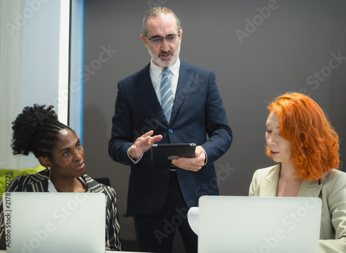 Photography senior male executive standing and holding digital tablet while guiding his smiling diverse female team members use laptops in modern corporate boardroom
