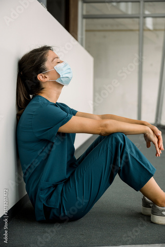 Exhausted nurse in scrubs and mask sitting on hospital floor