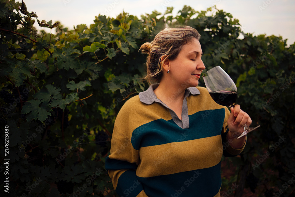 Fototapeta premium Woman enjoying red wine while standing among grapevines in a vineyard, wearing a striped sweater and holding a wine glass, with lush green foliage surrounding her