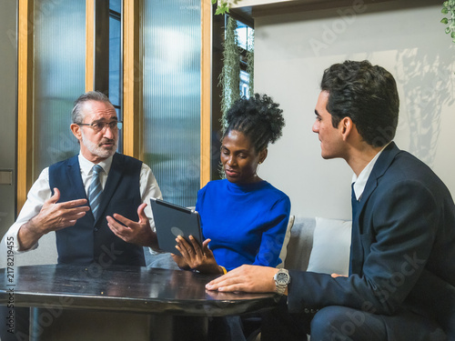 senior businessman explaining strategy with animated hand gestures to diverse multiethnic team of colleagues reviewing data on digital tablet in modern office meeting room.