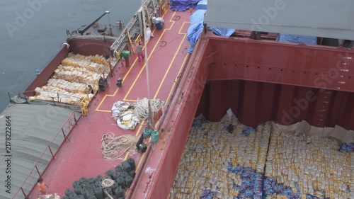 high angle view of a bulk carrier deck and open cargo hold loading bagged goods from an alongside barge with slings, rigging ropes, and manual activities 