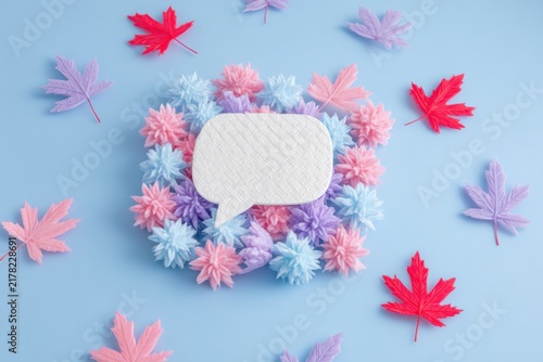 Colorful paper flowers and maple leaves surround a white speech bubble on a pastel blue background, creating a vibrant and artistic composition.