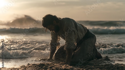 A man kneels on a sandy beach by the ocean as waves wash over him during sunset