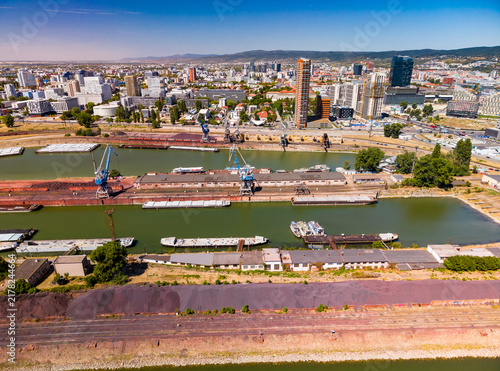View of a busy harbor area with boats and buildings under a clear sky. Boats are docked at a harbor surrounded by urban buildings and green areas on a sunny day.