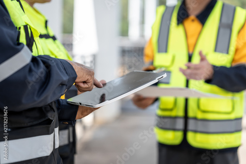 Close-up of construction workers in high visibility vests using a tablet and clipboard at an indoor work site, teamwork, inspection and project planning.