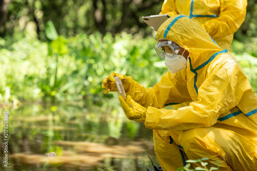 Environmental health workers in yellow protective suits collect a water sample from a stream in a green forest area.