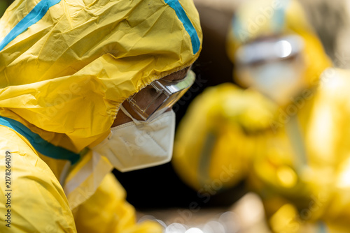 Close-up of a healthcare worker in yellow protective suit, goggles and face mask, with another hazmat worker blurred in the background.