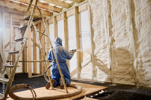 Worker kneeling and spraying polyurethane foam insulation into wall cavity of timber frame house