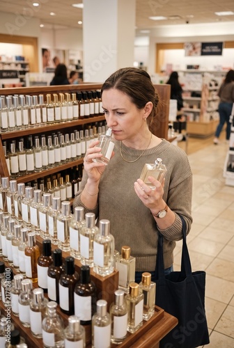 A woman stands in a store looking at a selection of perfume bottles. She holds a bottle up to her nose, taking in the scent. Customers are browsing nearby