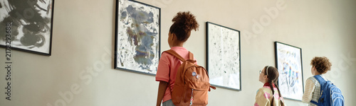 Diverse children with backpacks looking at paintings on wall in art gallery, rear view. Image suitable for school field trip promotions and cultural education campaigns