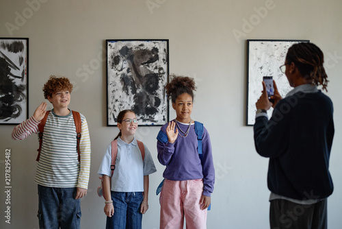 Diverse group of children posing for photo in art gallery while African American teacher taking picture with smartphone, promoting school field trips and cultural education
