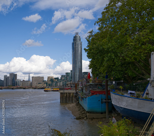 Photography Colorful residential houseboats moored along the River Thames in London, England, with modern high-rise buildings and the city skyline visible in the background